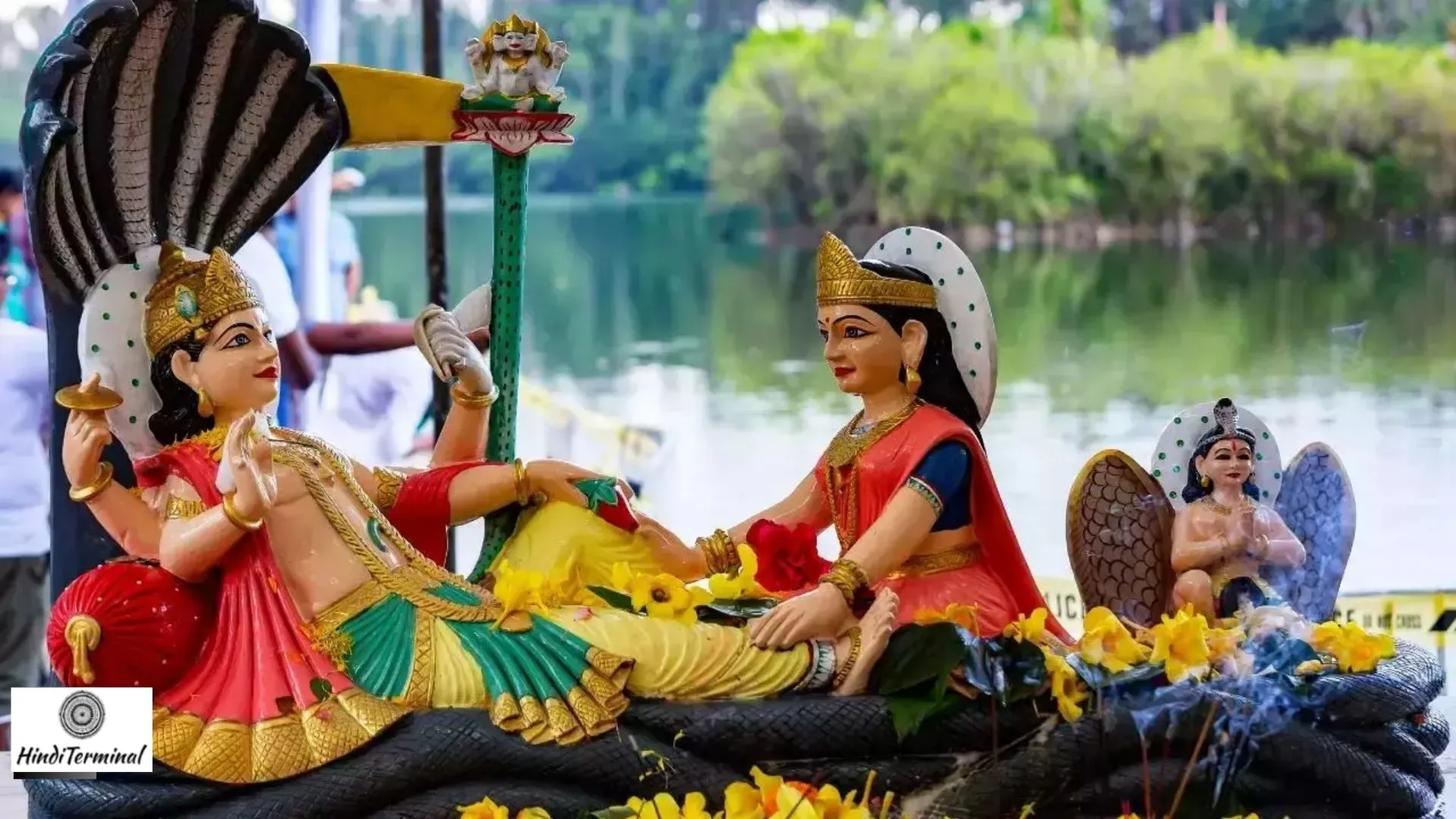 A vibrant idol of Lord Vishnu resting on Sheshnag with Goddess Lakshmi by his side, adorned with flowers, placed near a serene lake.